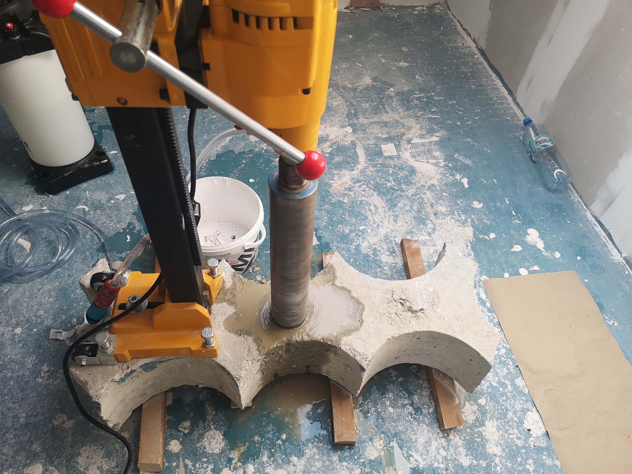 Żółta wiertnica rdzeniowa w trakcie wiercenia otworów w betonowym elemencie o kształcie łuków, z widocznym chłodziwem i fragmentami odpadów na podłodze.