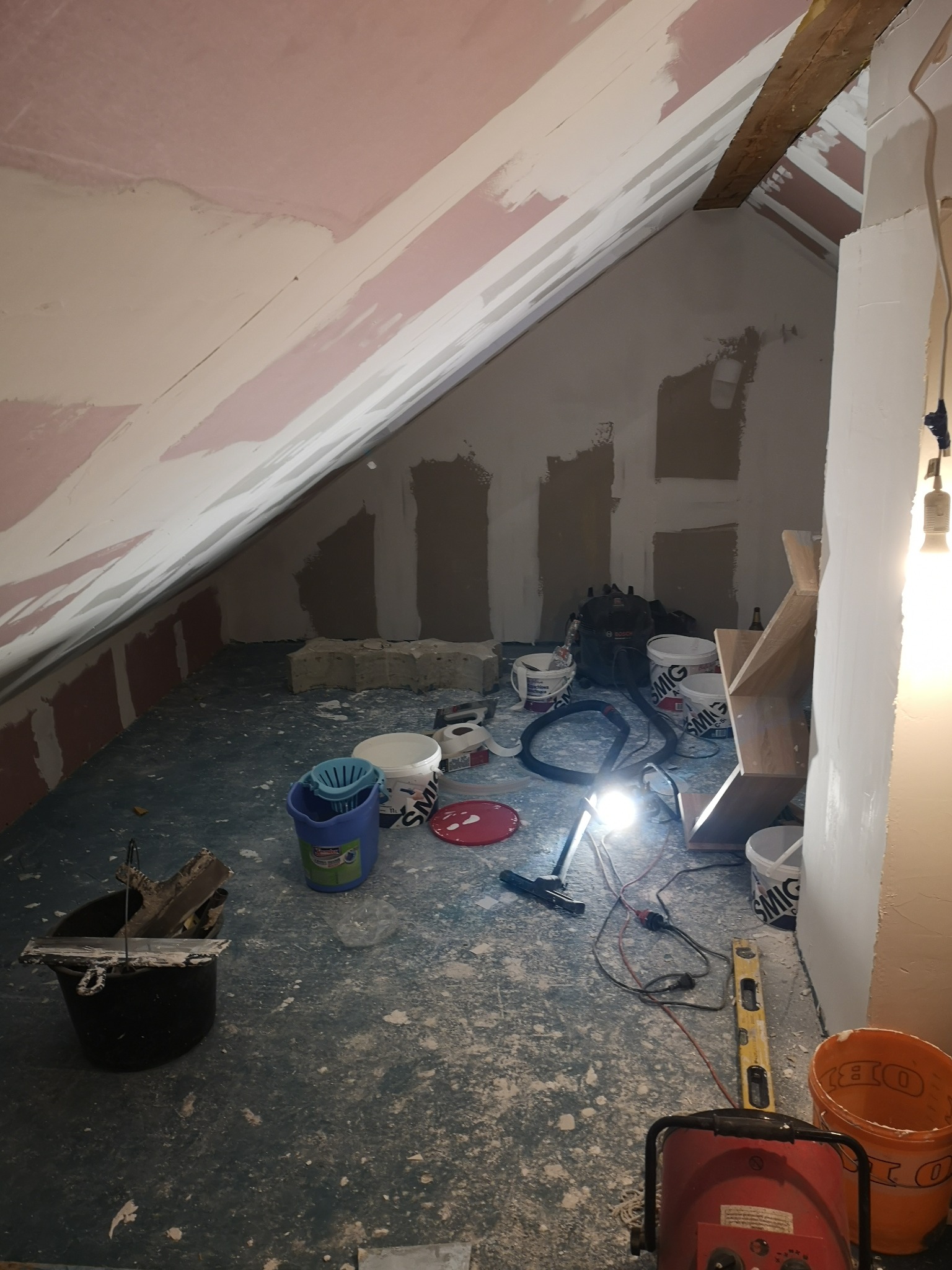 Poddasze w trakcie prac wykończeniowych: ściany pokryte wstępną warstwą, widoczne wiadra z farbą, szpachle, poziomica i lampa robocza.