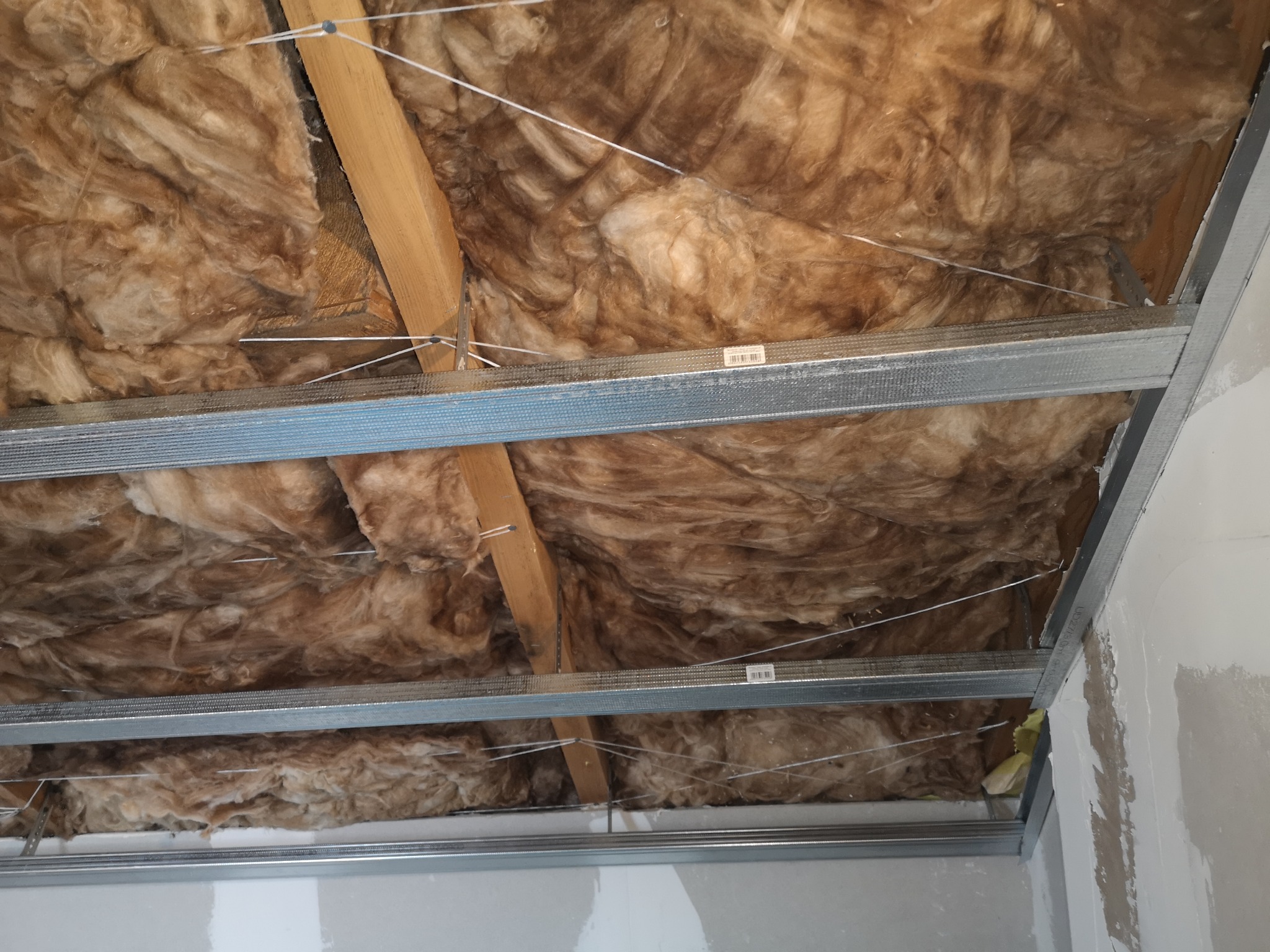 Izolacja z wełny mineralnej zamontowana pod sufitem podwieszanym z metalową konstrukcją, widoczne drewniane elementy konstrukcyjne.