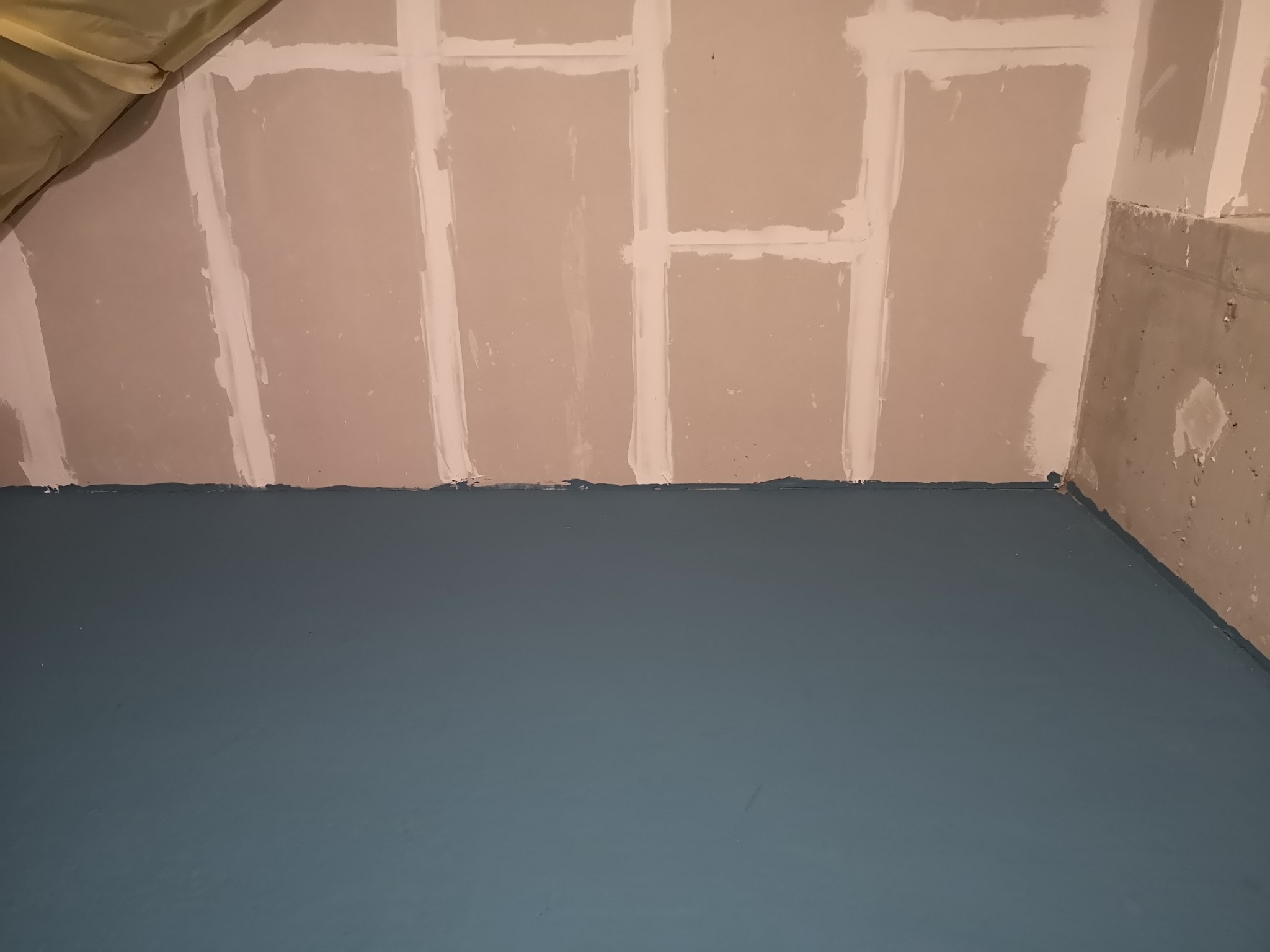 Przygotowane do malowania ściany z płyt gipsowo-kartonowych, zabezpieczone taśmą łączenia, z pomalowaną na niebiesko podłogą, widok na narożnik pokoju.