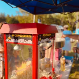 Automat do popcornu z napisem 'Popcorn' i rysunkiem postaci, wypełniony świeżym popcornem, ustawiony pod niebieskim namiotem na tle zieleni, z widocznymi osobami w tle.