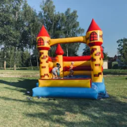 Dzieci bawią się w dmuchanym zamku w kształcie fortecy z żółto-czerwonymi wieżami na trawiastym terenie w słoneczny dzień.