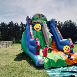 Watę Lizać - Duży, kolorowy dmuchany zamek ze zjeżdżalnią na trawie, z bawiącymi się dziećmi, nazwa zamku Lemurs z motywem papugi na górze.
