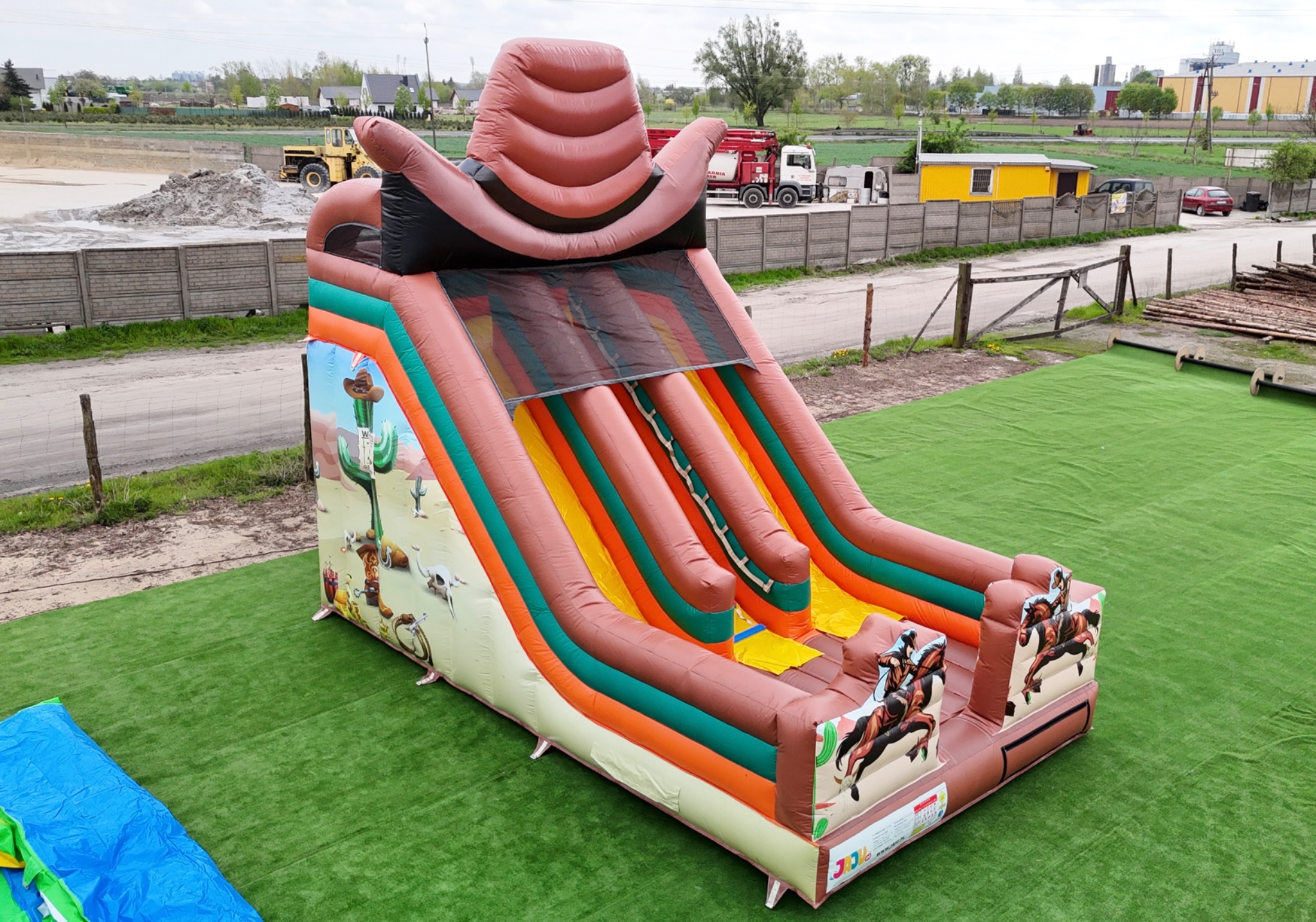 Duży dmuchany zamek w stylu kowbojskim z motywami kaktusów, koni i kapeluszy, ustawiony na sztucznej trawie na zewnątrz.