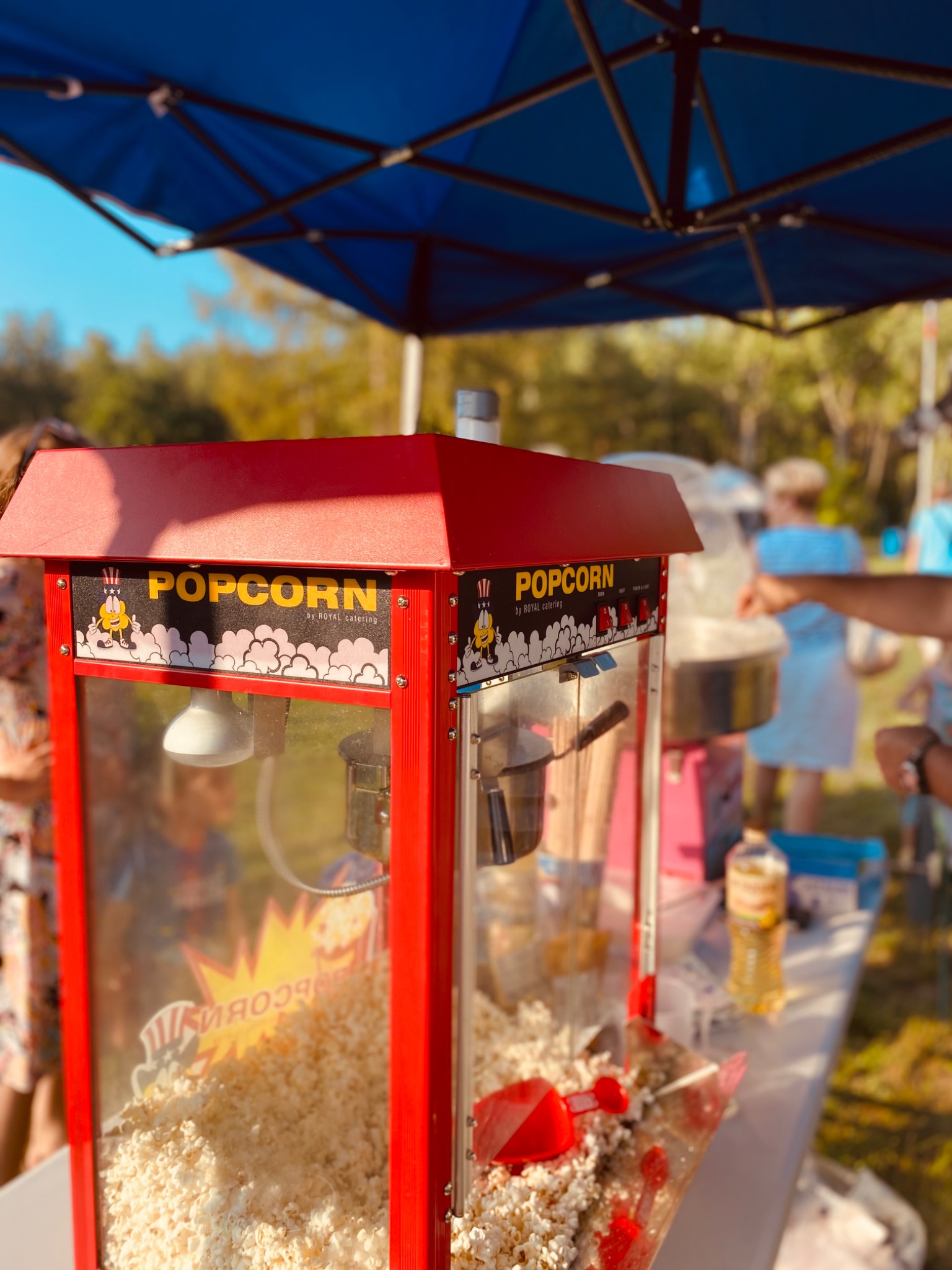 Automat do popcornu z napisem 'Popcorn' i rysunkiem postaci, wypełniony świeżym popcornem, ustawiony pod niebieskim namiotem na tle zieleni, z widocznymi osobami w tle.