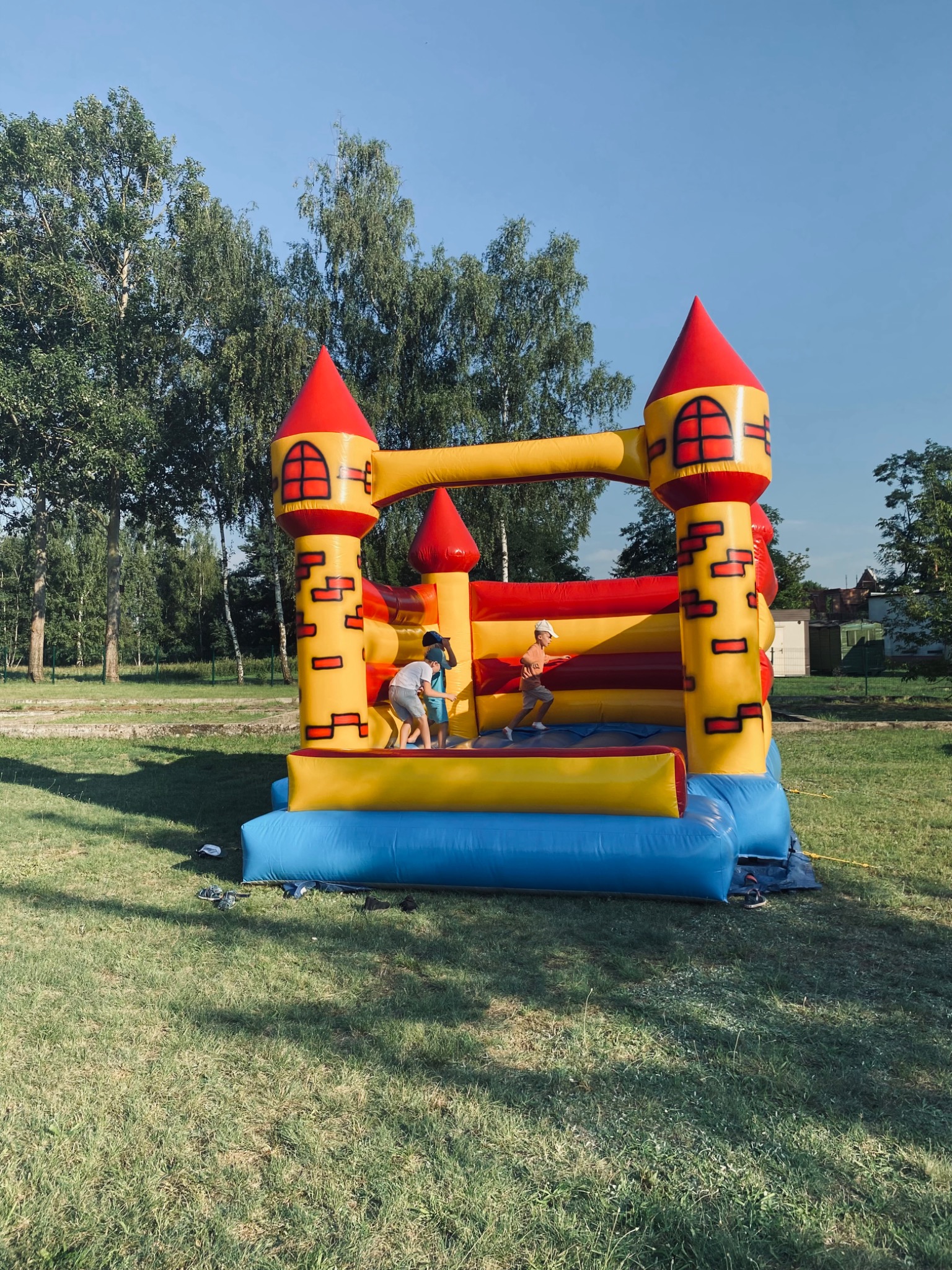 Dzieci bawią się w dmuchanym zamku w kształcie fortecy z żółto-czerwonymi wieżami na trawiastym terenie w słoneczny dzień.