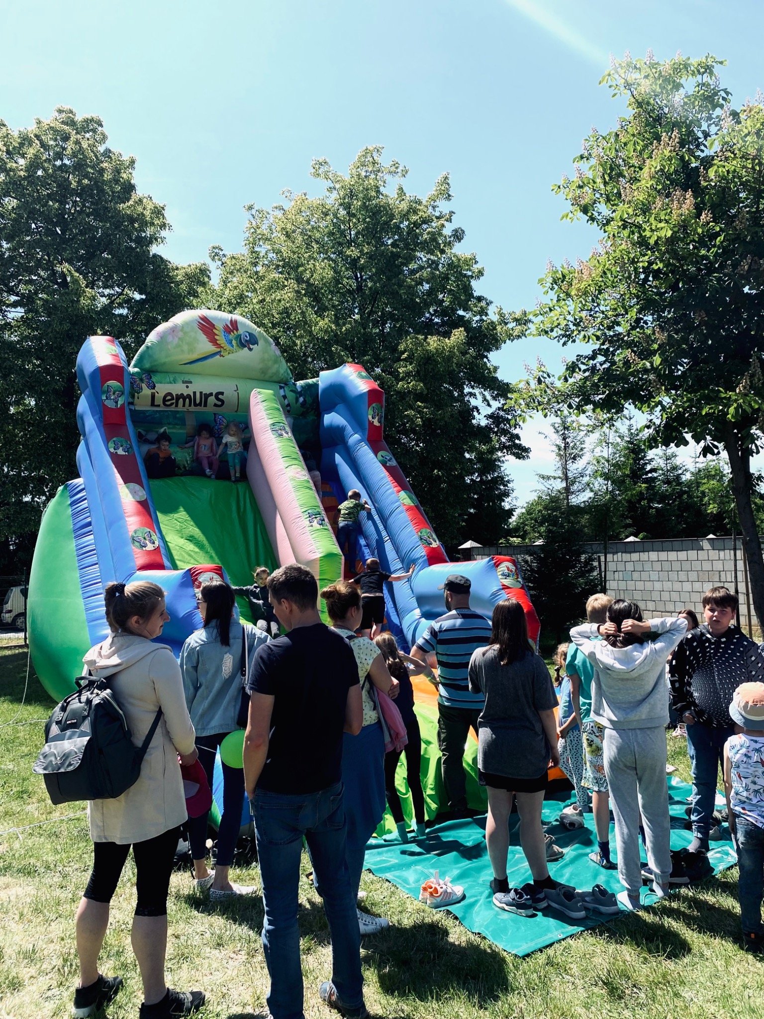 Dmuchana zjeżdżalnia z motywem dżungli i napisem 'Lemurs' otoczona grupą dzieci i dorosłych na trawiastym terenie w słoneczny dzień.