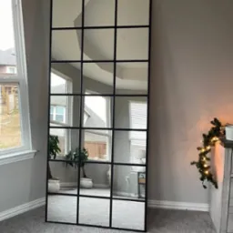 Wysokie lustro z czarnymi szprosami imitujące drzwi, odbijające wnętrze pokoju z oknem i roślinami doniczkowymi.