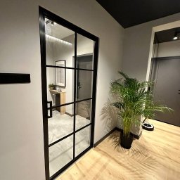 DomLoftu - Nowoczesne drzwi wewnętrzne z czarną ramą i szklanymi panelami, widok na przedpokój z konsolą i obrazem, parkiet w odcieniach beżu, roślina doniczkowa i robot sprzątający w tle.