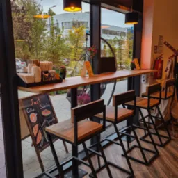 Wnętrze kawiarni z wysokimi krzesłami barowymi o metalowej konstrukcji i drewnianych siedziskach przy długim drewnianym blacie pod oknem, widok na ulicę z drzewami i budynkiem w tle, tablica z menu...