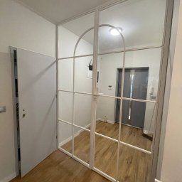 DomLoftu - Białe, szklane drzwi wewnętrzne z metalową ramą, podzielone na prostokątne i łukowate sekcje, oddzielające przedpokój z drewnianą podłogą od dalszej części mieszkania.