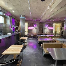 DomLoftu - Wnętrze lokalu gastronomicznego z surowymi ścianami z cegły, oświetlone neonowymi napisami i fioletowym światłem, z metalowymi i drewnianymi stolikami oraz krzesłami.