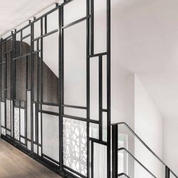 DomLoftu - Nowoczesna balustrada schodowa z czarną metalową ramą i wypełnieniami z różnych rodzajów szkła, w tym szkła ornamentowego i przezroczystego, zamocowana do sufitu, widok z dołu schodów.