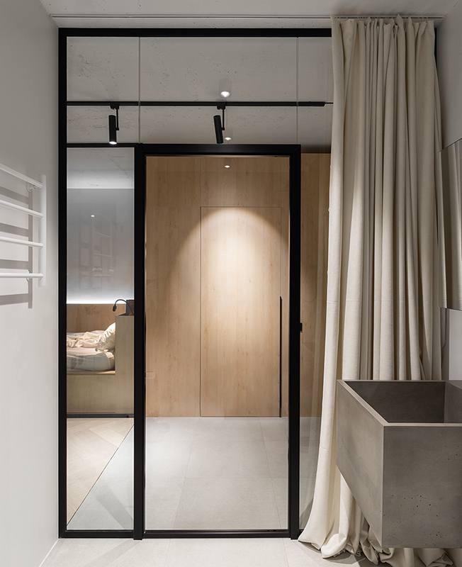 Nowoczesne, minimalistyczne wnętrze z czarną ramą szklanych drzwi, widok na sypialnię z drewnianą zabudową, betonowa umywalka i beżowe zasłony.
