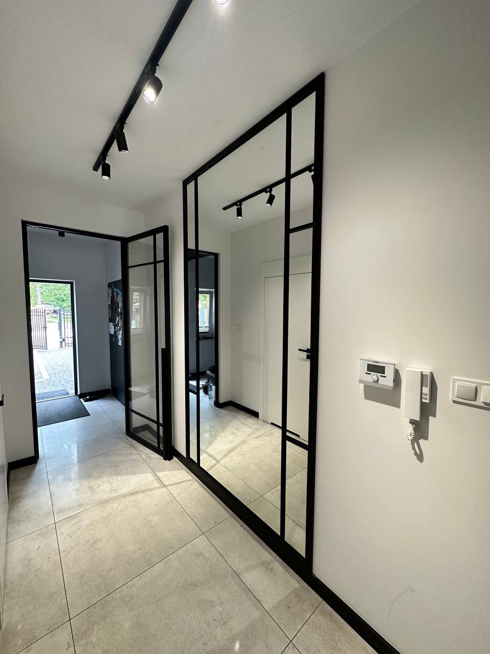 Nowoczesne wnętrze przedpokoju z szklaną ścianą działową w czarnej ramie, odzwierciedlającą oświetlenie sufitowe i dalszą część korytarza, z widocznym fragmentem wejścia do domu.