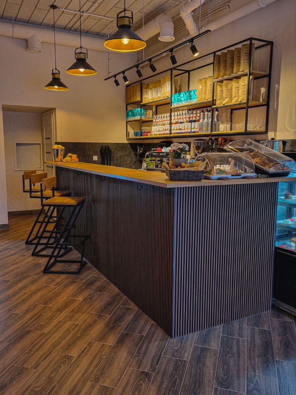 Bar w kawiarni z drewnianymi krzesłami barowymi, pionowo żłobkowanym panelem frontowym baru, półkami z kawą i napojami w tle, oświetlony wiszącymi lampami z czarnymi kloszami.