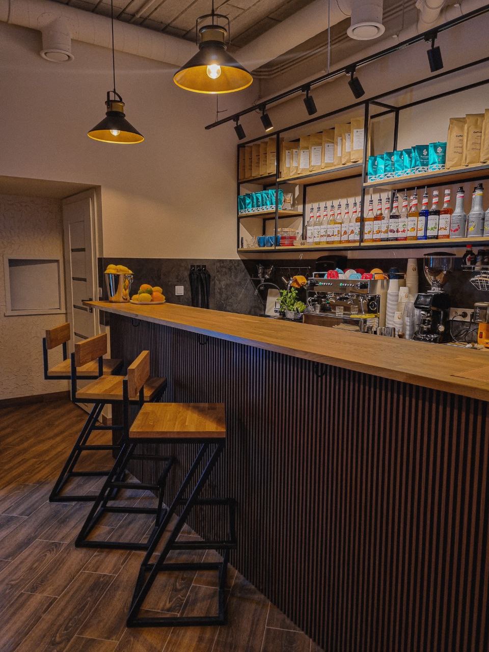 Wnętrze kawiarni z drewnianym blatem, metalowymi hokerami, półkami z kawą i syropami, oraz lampami w stylu industrialnym.