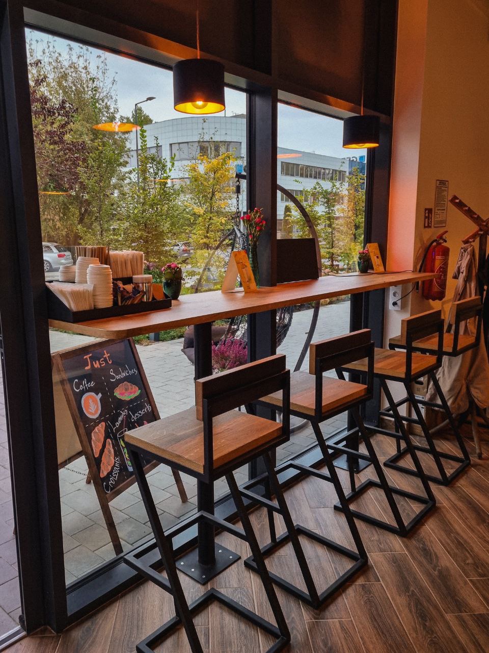 Wnętrze kawiarni z wysokimi krzesłami barowymi o metalowej konstrukcji i drewnianych siedziskach przy długim drewnianym blacie pod oknem, widok na ulicę z drzewami i budynkiem w tle, tablica z menu...