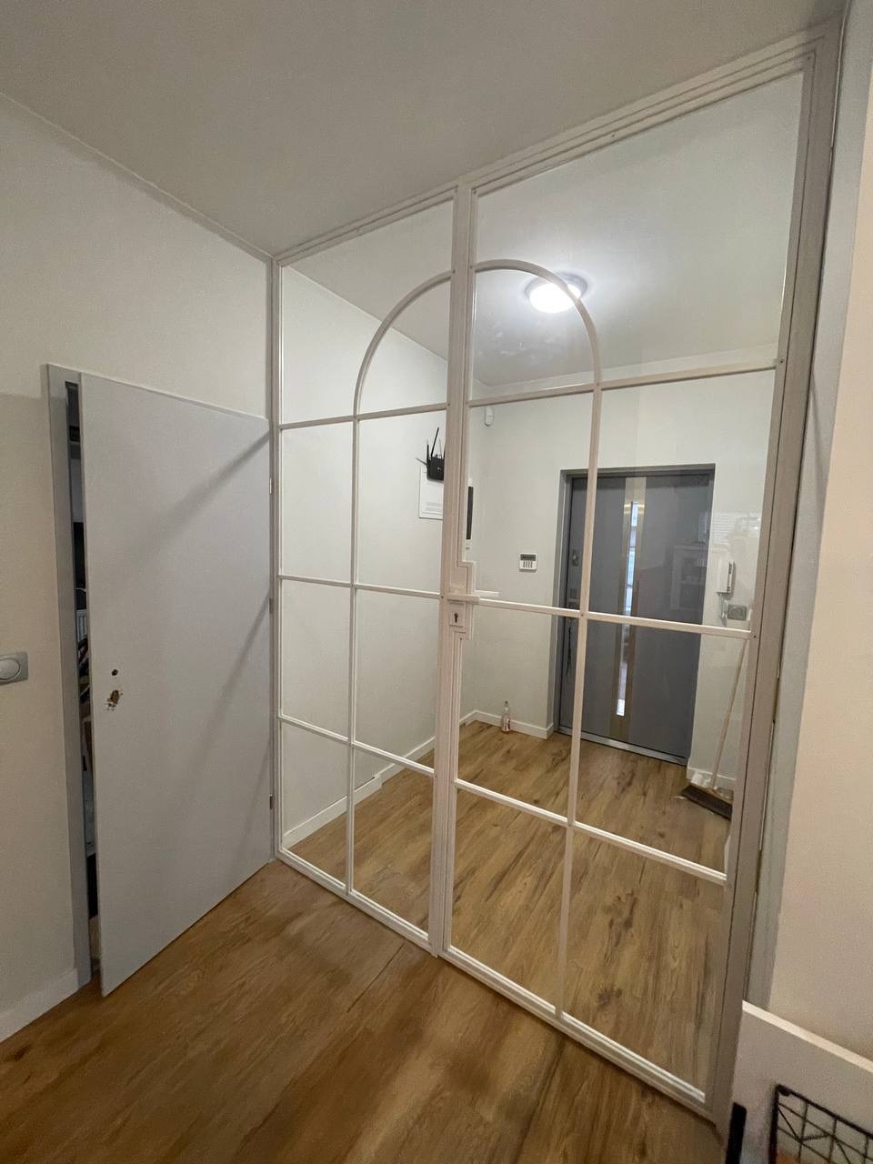 Białe, szklane drzwi wewnętrzne z metalową ramą, podzielone na prostokątne i łukowate sekcje, oddzielające przedpokój z drewnianą podłogą od dalszej części mieszkania.