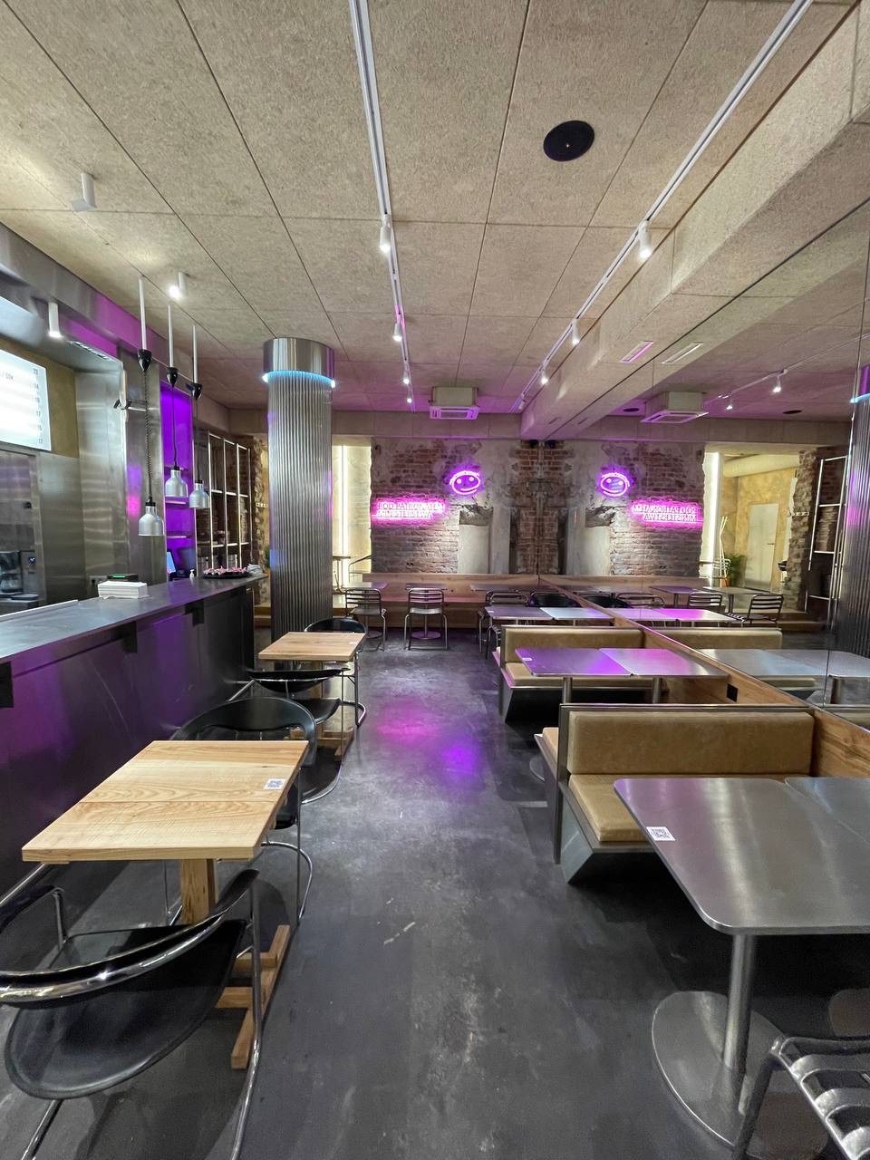 Wnętrze lokalu gastronomicznego z surowymi ścianami z cegły, oświetlone neonowymi napisami i fioletowym światłem, z metalowymi i drewnianymi stolikami oraz krzesłami.