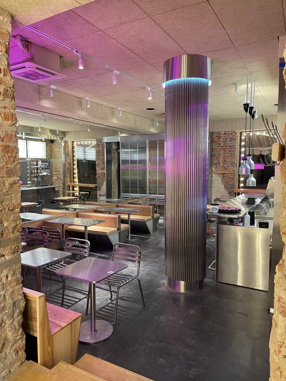 Wnętrze lokalu gastronomicznego z metalową kolumną podświetloną na fioletowo, widoczne stoliki, krzesła i boksy ze skórzanymi siedziskami, surowa cegła na ścianach.