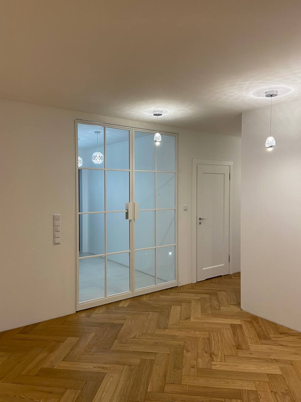 Nowoczesne, dwuskrzydłowe drzwi przesuwne szklane z białymi szprosami, zamontowane w jasnym wnętrzu z parkietem w jodełkę i minimalistycznym oświetleniem.