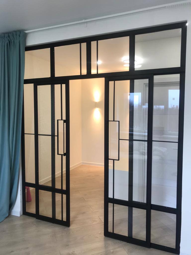 Przesuwne drzwi w stylu loft z czarnymi szprosami, dzielące przestrzeń, widoczne fragmenty parkietu i zasłony w tle.