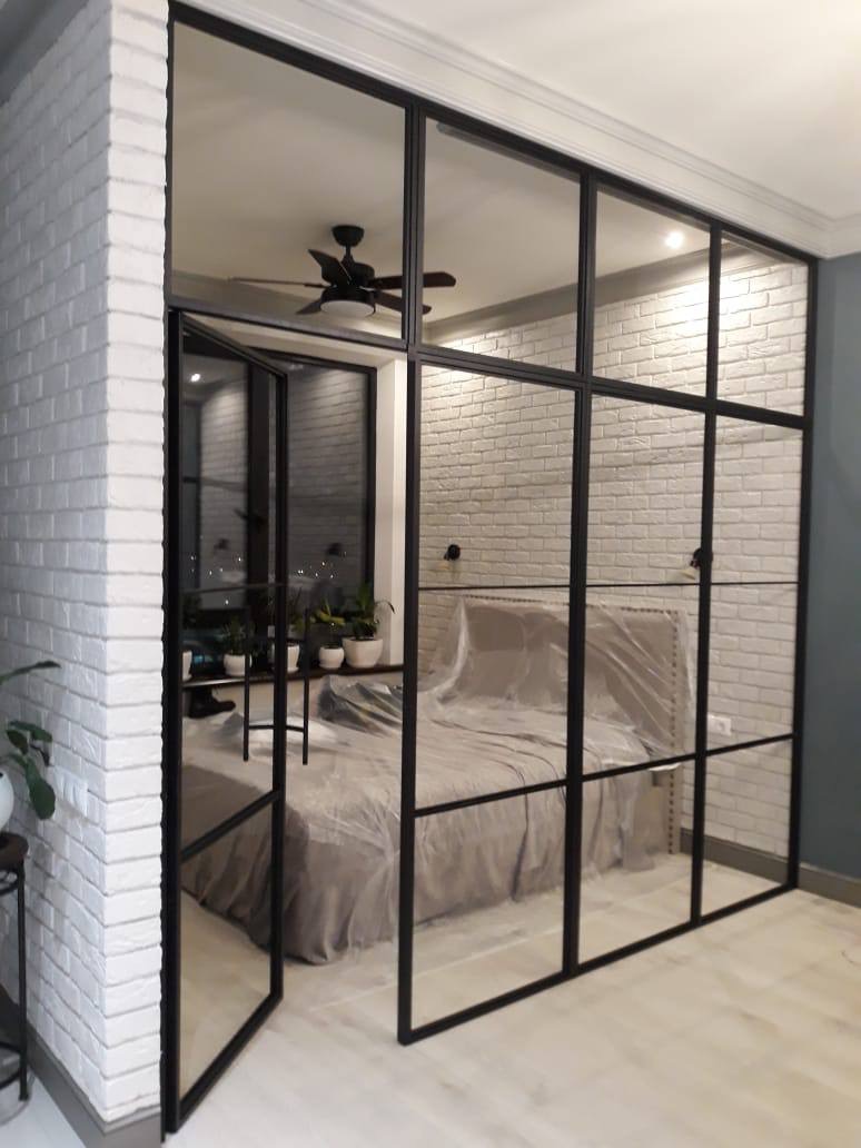 Szklane drzwi przesuwne w stylu loft z czarnymi szprosami oddzielają sypialnię z ceglaną ścianą i wentylatorem sufitowym, widoczne częściowo otwarte skrzydło.