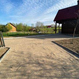 FUH Roboty Ziemne i Wyburzenia Jakub Żuchowski - Utwardzona, piaszczysta droga dojazdowa do domu z czerwonym dachem, obrzeża z kostki, błękitne niebo z chmurami w tle. Widoczne budynki w oddali.