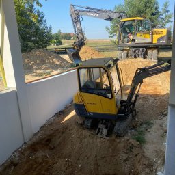 FUH Roboty Ziemne i Wyburzenia Jakub Żuchowski - Żółta mini koparka Volvo EC15B w wykopie obok białej ściany, w tle większa koparka Volvo EW160C i drzewa. Prace ziemne w słoneczny dzień.