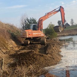 FUH Roboty Ziemne i Wyburzenia Jakub Żuchowski - Pomarańczowa koparka Atlas 1504 LC na brzegu stawu, nabiera ziemię. W tle widać drzewa i budynek. Praca w toku, ziemia częściowo w wodzie, odbicie koparki w tafli.