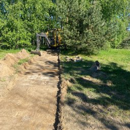 FUH Roboty Ziemne i Wyburzenia Jakub Żuchowski - Żółta minikoparka na działce, przygotowuje teren pod budowę. Wykopany pas ziemi, widoczne drzewa w tle. Prace ziemne w toku, słoneczny dzień.