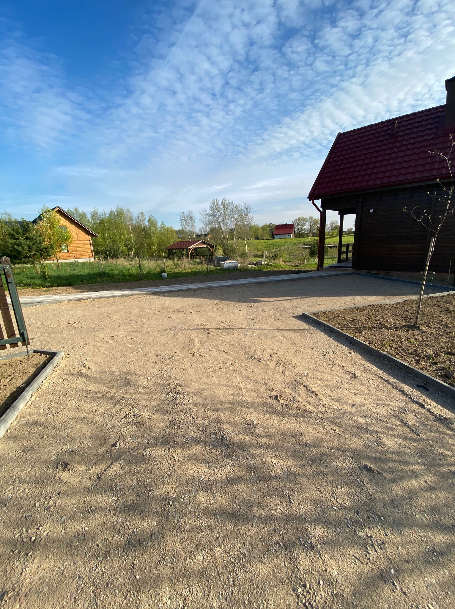 Utwardzona, piaszczysta droga dojazdowa do domu z czerwonym dachem, obrzeża z kostki, błękitne niebo z chmurami w tle. Widoczne budynki w oddali.