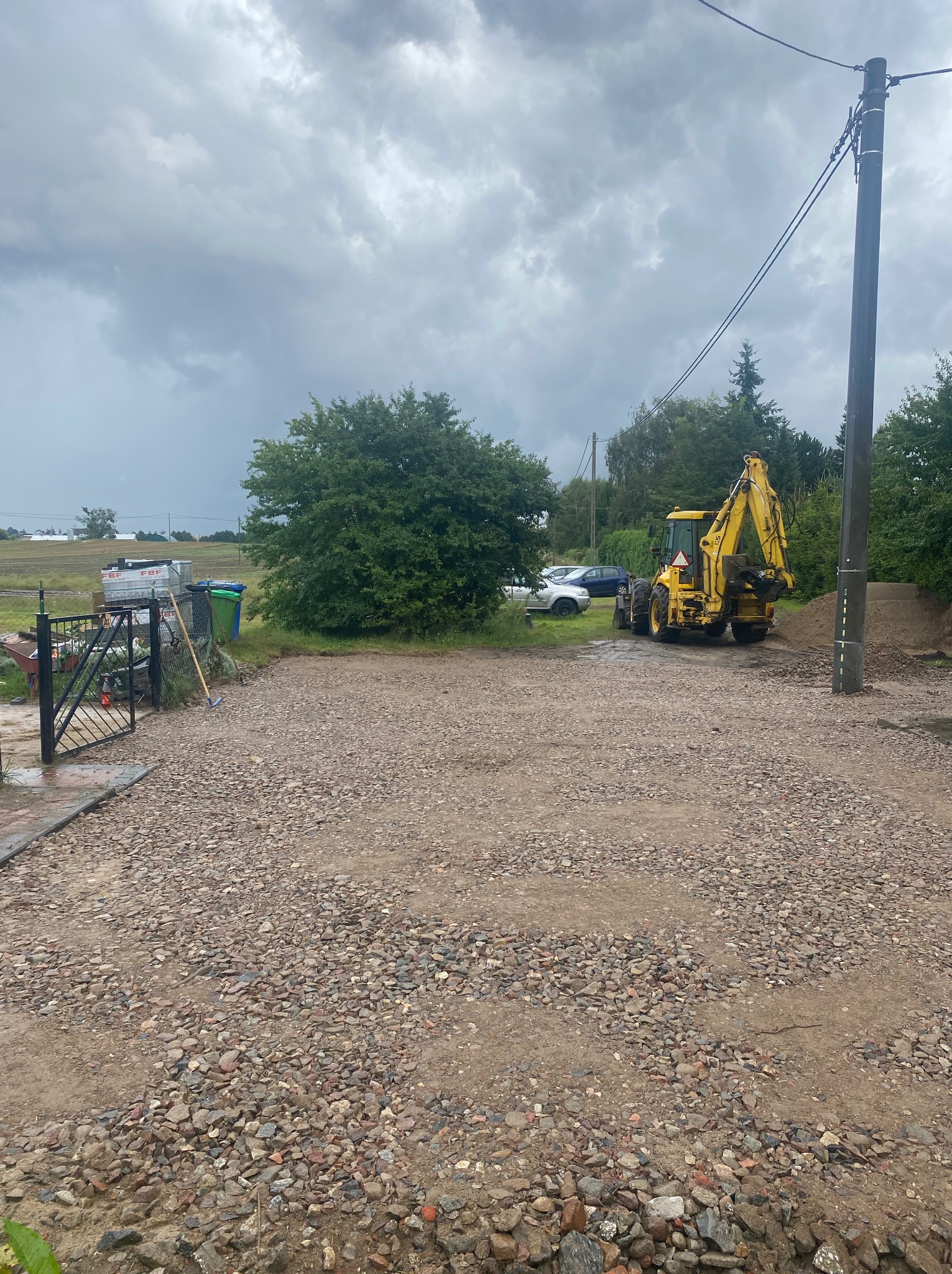 Żwirowa nawierzchnia z żółtą koparką w tle, częściowo zasłoniętą przez usypaną ziemię. Widoczne drzewa, słup elektryczny i pochmurne niebo.