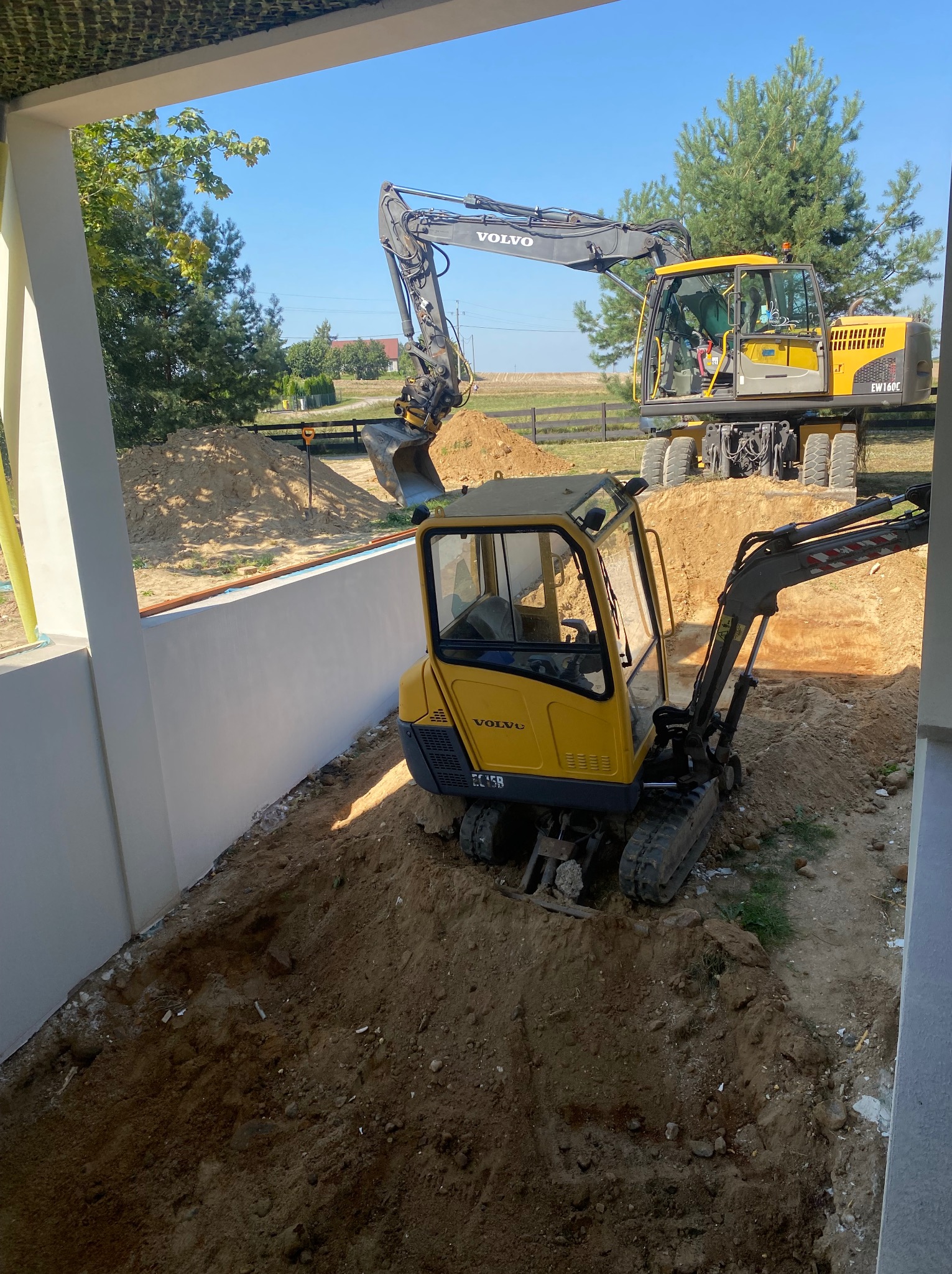 Żółta mini koparka Volvo EC15B w wykopie obok białej ściany, w tle większa koparka Volvo EW160C i drzewa. Prace ziemne w słoneczny dzień.