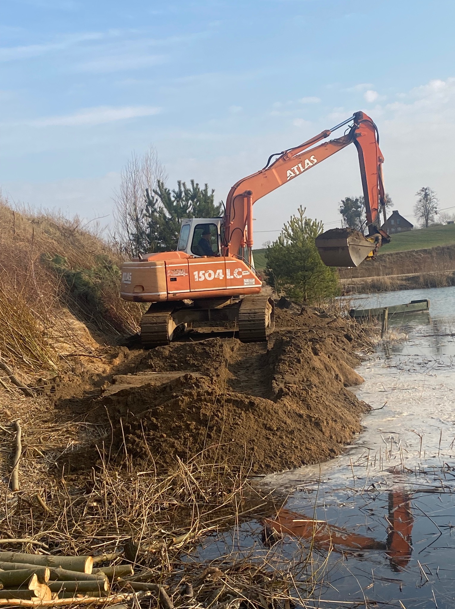 Pomarańczowa koparka Atlas 1504 LC na brzegu stawu, nabiera ziemię. W tle widać drzewa i budynek. Praca w toku, ziemia częściowo w wodzie, odbicie koparki w tafli.