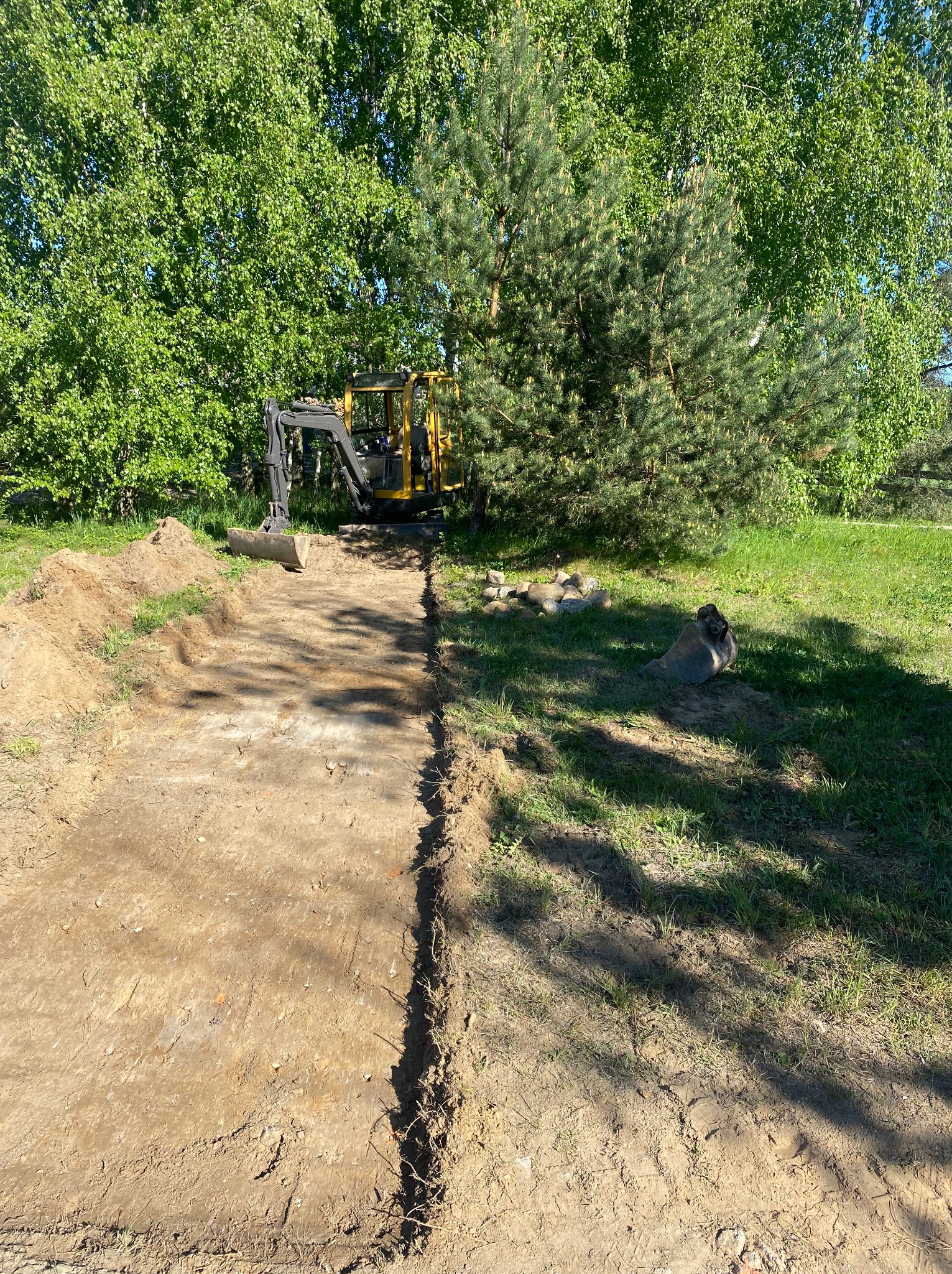 Żółta minikoparka na działce, przygotowuje teren pod budowę. Wykopany pas ziemi, widoczne drzewa w tle. Prace ziemne w toku, słoneczny dzień.