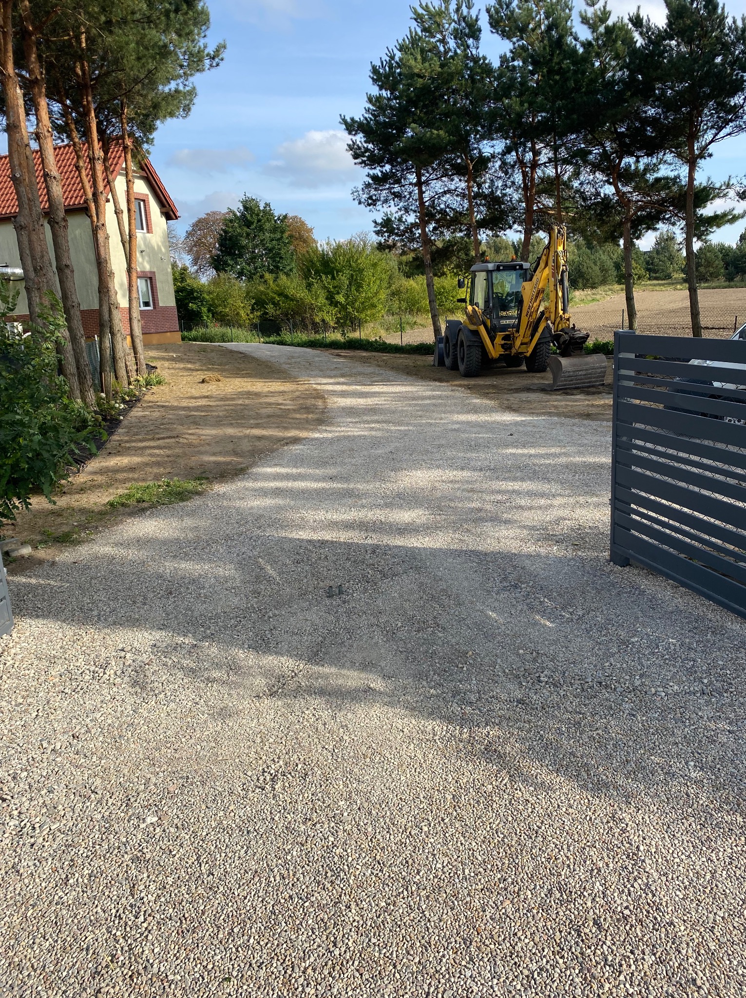 Żwirowa droga do domu z żółtą koparką w tle, widok zza szarej bramy. Drzewa wzdłuż drogi i błękitne niebo. Utwardzona nawierzchnia.