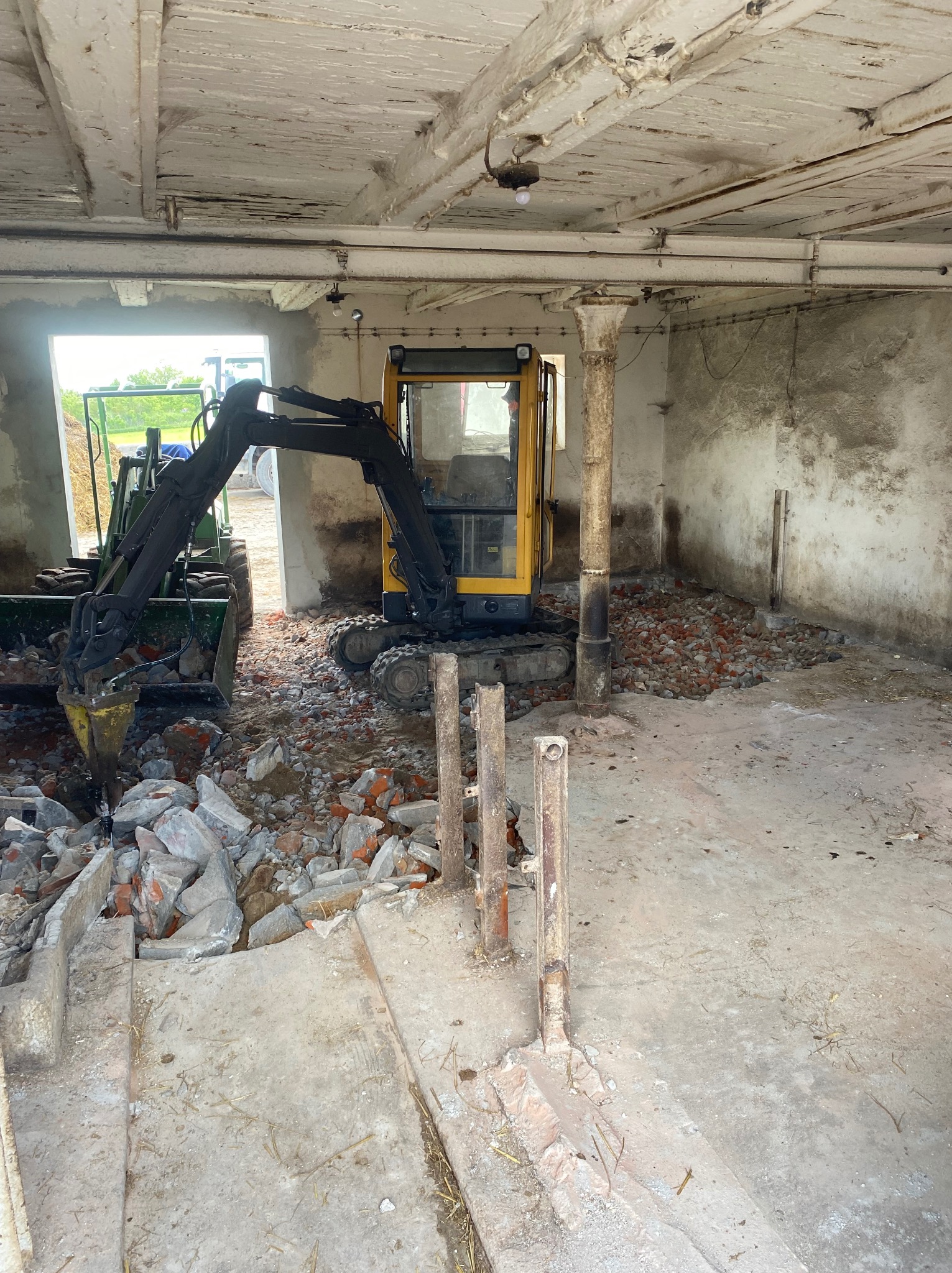 Wnętrze budynku w trakcie rozbiórki: mini koparka z młotem pneumatycznym kruszy gruz ceglany. Widoczne fragmenty ścian i stropu. Otwarty przejazd na zewnątrz.