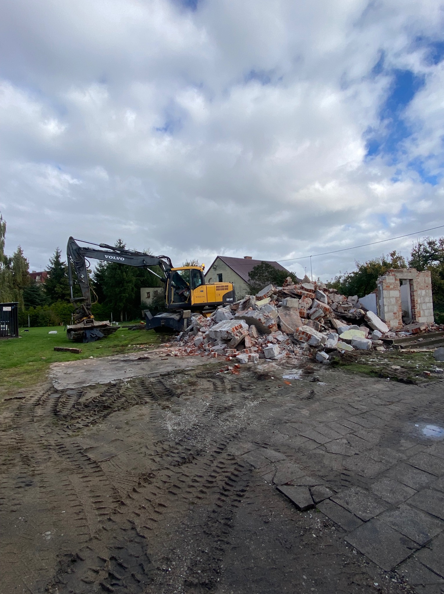 Żółta koparka Volvo przy stercie gruzu po rozbiórce w Biskupcu. Widoczne fragmenty muru i ślady opon na ziemi. Prace rozbiórkowe w toku, pochmurne niebo.