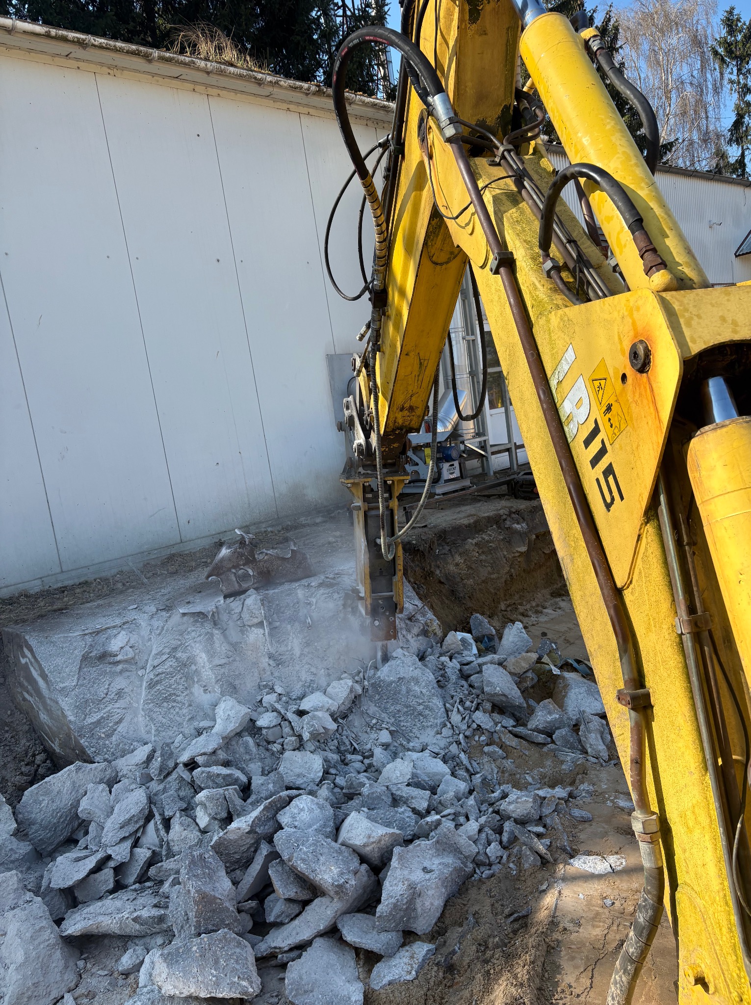 Żółty młot hydrauliczny kruszy szary kamień na placu budowy. Widoczne fragmenty maszyny, gruz i biała ściana budynku w tle. Ujęcie z bliska, dynamiczne.