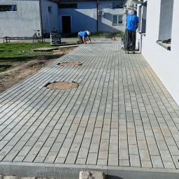 K.P. Digger - Układanie kostki brukowej na posesji przy budynku. Widoczne studzienki rewizyjne, pracownicy w trakcie pracy i sprzęt budowlany. Jasne, słoneczne oświetlenie.