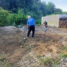 K.P. Digger - Prace ziemne w Barwicach: Dwóch mężczyzn wyrównuje teren po wyburzeniu, przygotowując go pod dalsze prace budowlane. W tle sterta drewna i zieleń.