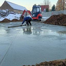 K.P. Digger - Pracownik wyrównuje wylewkę betonową na fundamencie. W tle koparka, hałda ziemi i ośnieżony budynek. Widoczne ślady zacierania betonu.