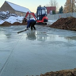 K.P. Digger - Wyrównywanie wylewki betonowej w wykopie pod fundamenty, na budowie stoi mała koparka, w tle budynek i ogrodzenie. Praca w toku, widoczne narzędzia i materiały.