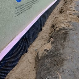 K.P. Digger - Izolacja fundamentu budynku z użyciem folii kubełkowej i styropianu, zasypana piaskiem. Widoczny fragment ściany z tekstem informacyjnym.