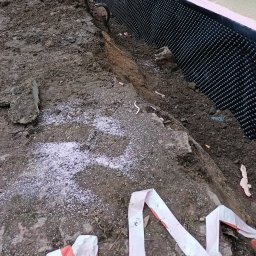 K.P. Digger - Fundament zabezpieczony folią kubełkową, widoczny wykop z ziemią i biały proszek rozsypany wokół. Taśma ostrzegawcza leży na ziemi.