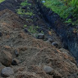 K.P. Digger - Wykopany rów w ziemi, z hałdą ziemi obok, częściowo porośnięty zielenią. Widoczne odciski opon w wykopie. Kamienie w ziemi.