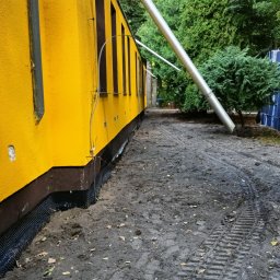 K.P. Digger - Żółty budynek z izolacją fundamentów i śladami opon w ziemi. Widoczna instalacja rury spustowej. W tle drzewa.