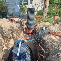 K.P. Digger - Instalacja zbiornika retencyjnego w wykopie, widoczne rury, studzienka i poziomica. W tle budynek mieszkalny i drzewa. Prace ziemne w toku.